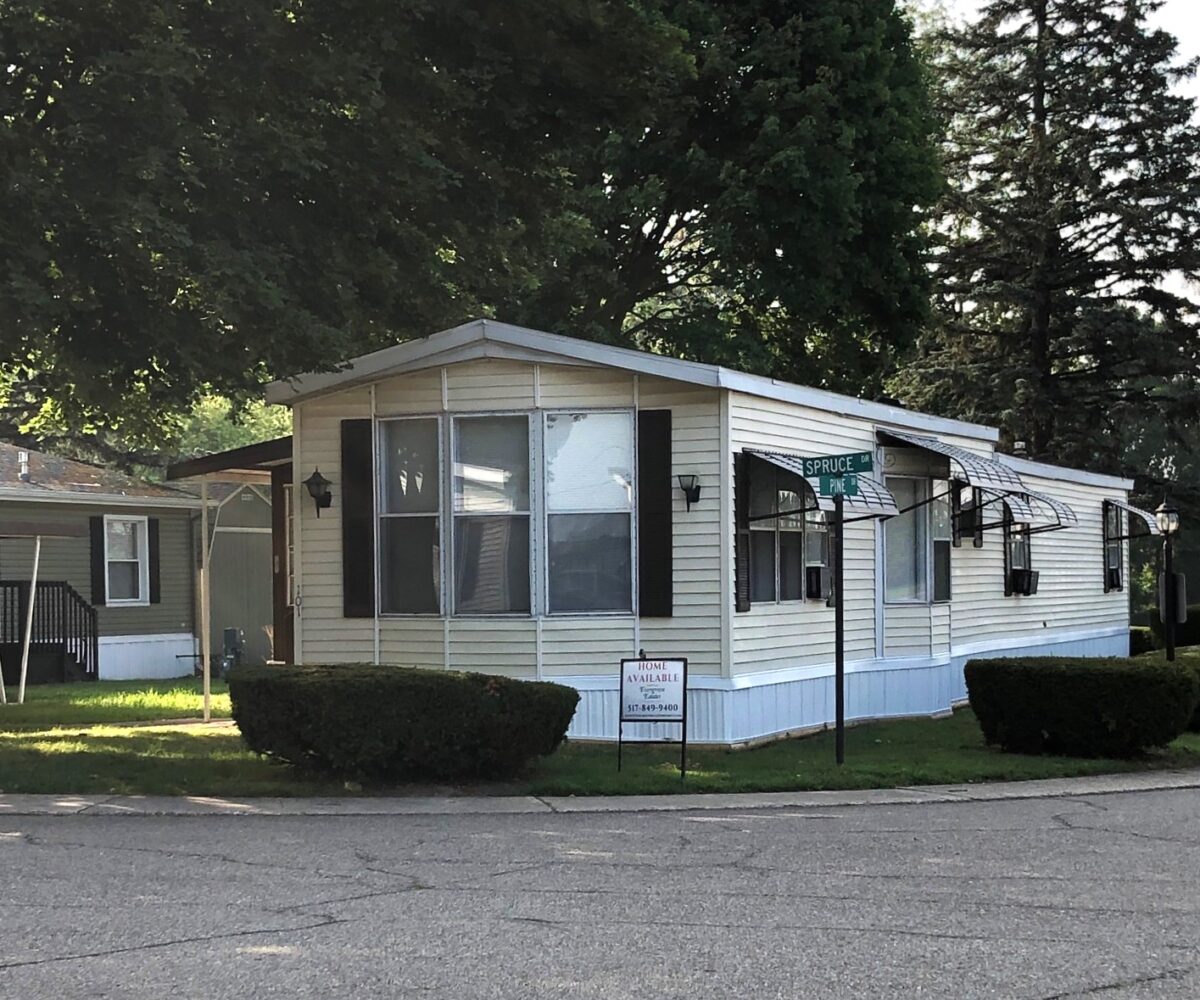 Single-story beige mobile home on a corner lot with awnings and a 'Home Available' sign in the yard.