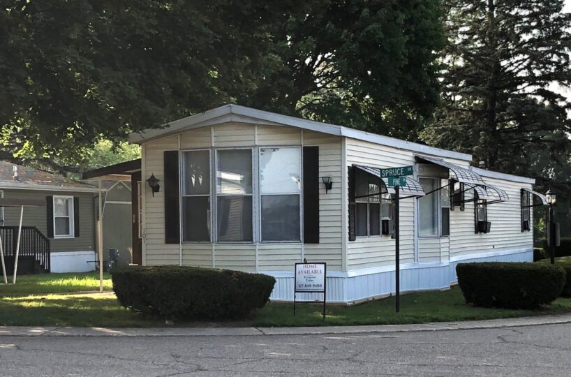 Single-story beige mobile home on a corner lot with awnings and a 'Home Available' sign in the yard.