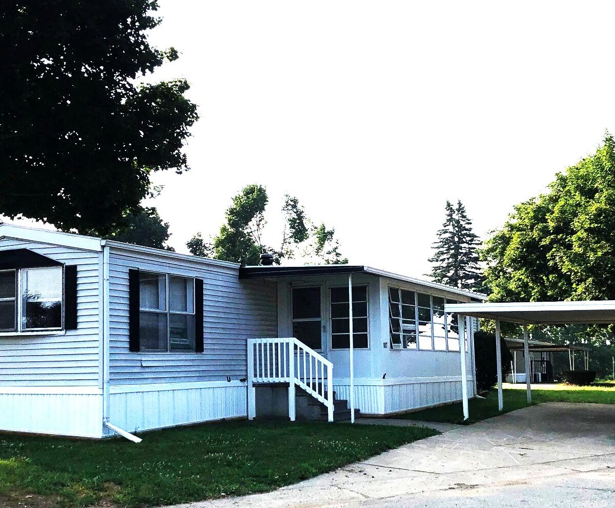 Light blue manufactured home with a small front porch, white railing, and carport on the right, set on a grassy lawn.
