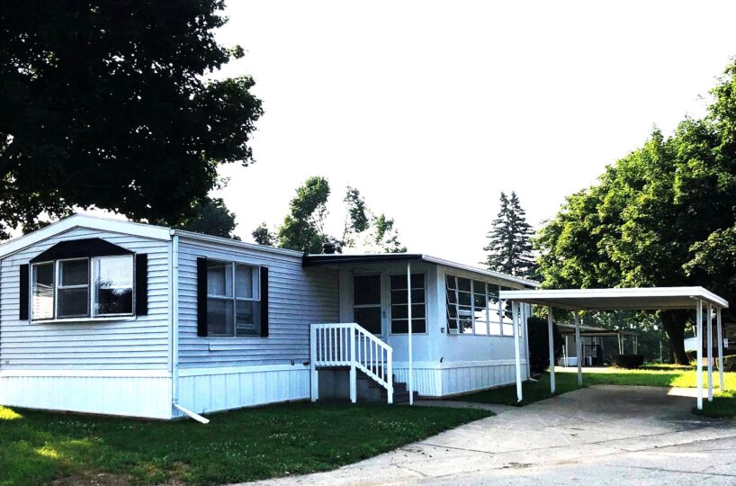 Light blue manufactured home with a small front porch, white railing, and carport on the right, set on a grassy lawn.