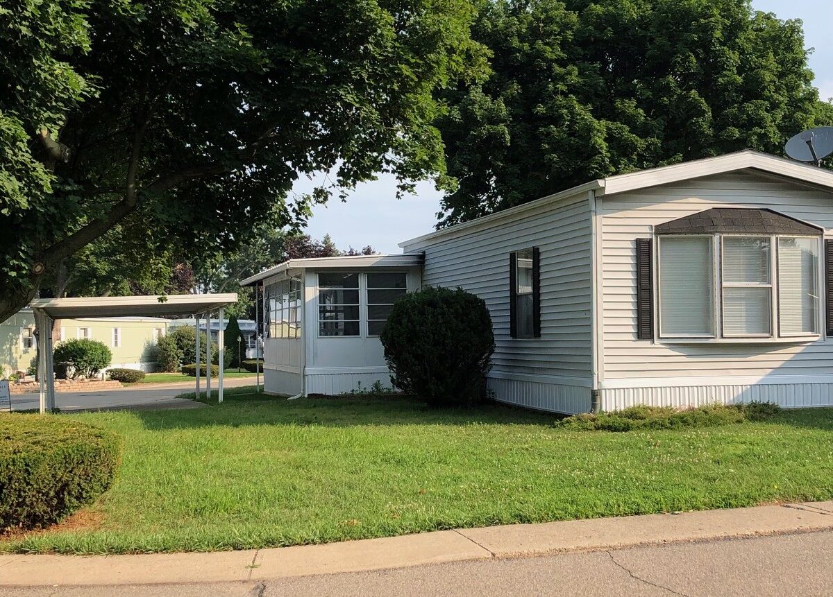 White manufactured home with a bay window and black shutters in a mobile home park, large tree shading the lawn in the foreground.