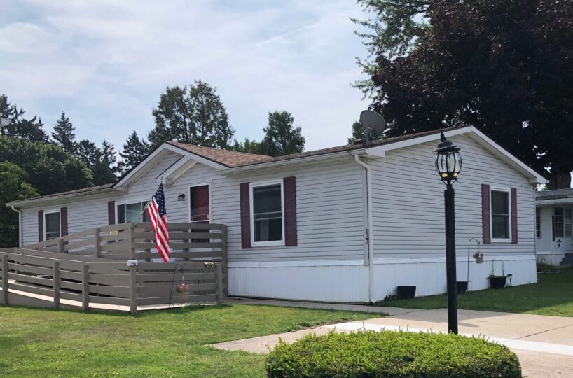 Single-story white mobile home with a wooden fenced deck, US flag, and a lamp post in the front yard.